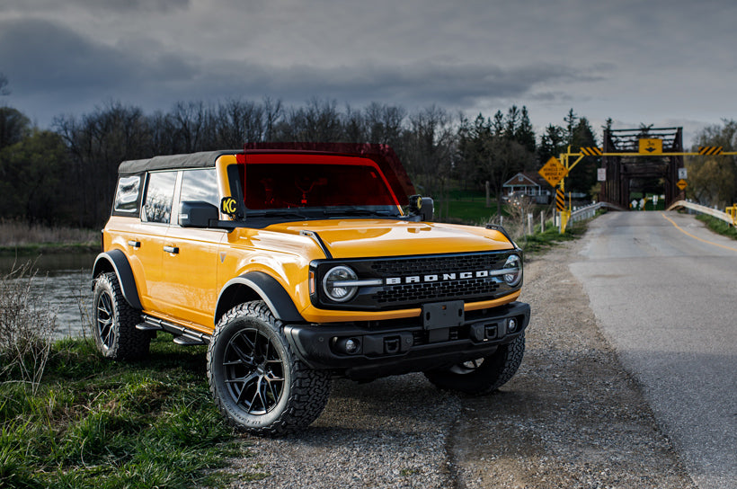 A yellow Ford Bronco with a protective film on the windshield, parked on the side of a road. With a bridge in the background.