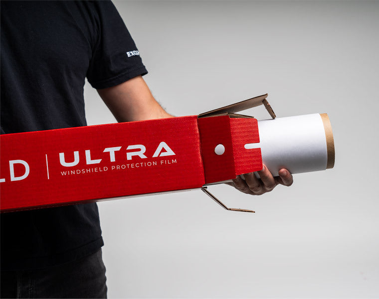 Person holding a role of ExoShield ULTRA WPF (windshield protection film kit) in their right hands. Standing in front of a white backdrop.