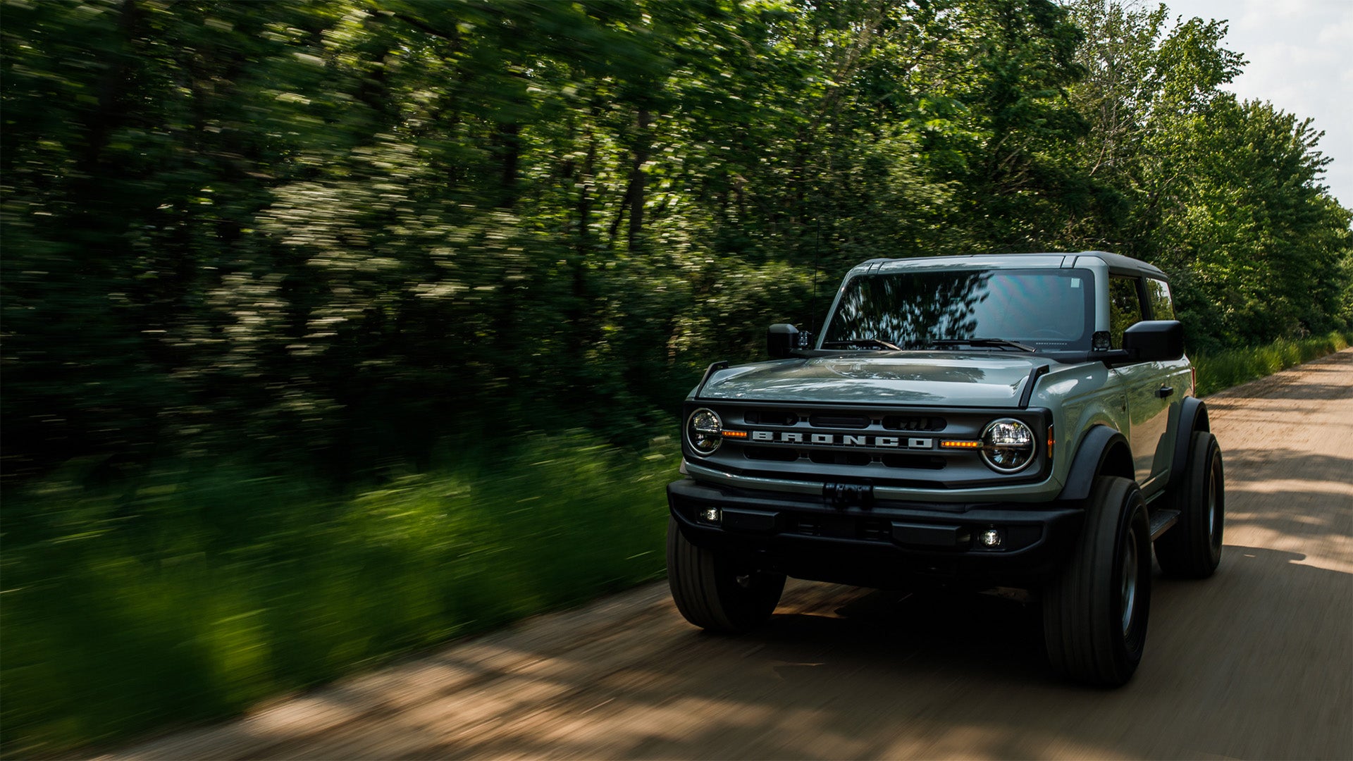 A dark gray Ford Bronco SUV driving on a dirt road surrounded by a blurred green forest.
