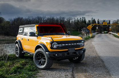 A yellow Ford Bronco with a protective film on the windshield, parked on the side of a road. With a bridge in the background. 