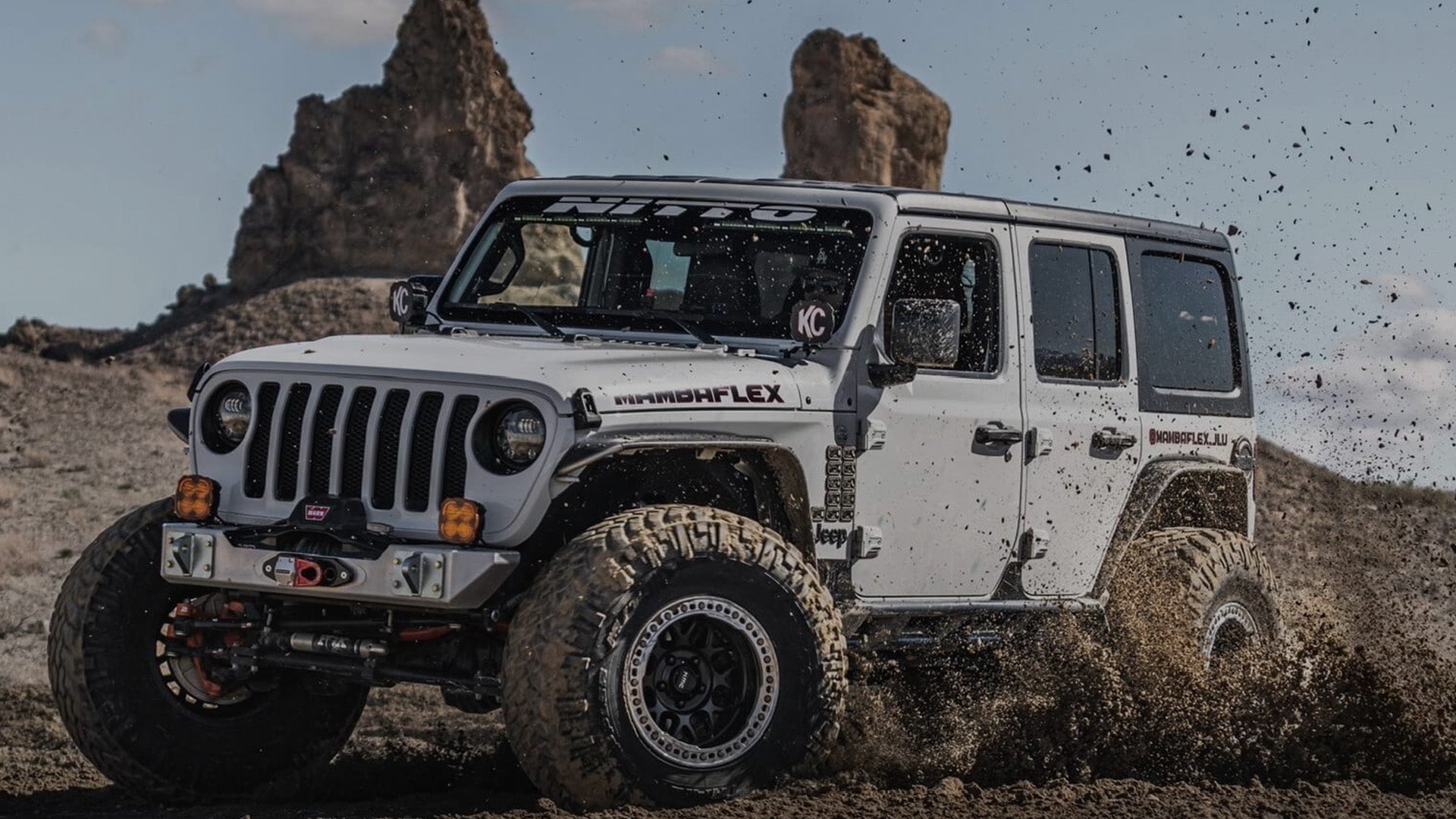 A white custom off-road Jeep Wrangler with large knobby tires and a front bumper brush guard, driving through mud in a desert landscape with rock formations in the background.