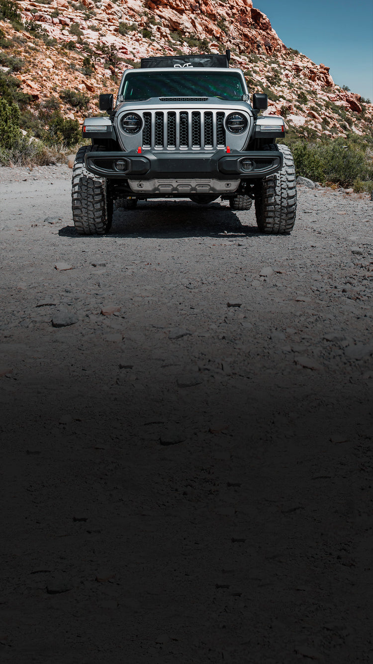 A black Jeep Wrangler parked on a dark dirt road with a rocky desert hillside in the background.