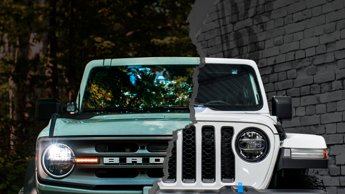 Two off-road vehicles, a green Ford Bronco and a white Jeep, parked side by side on a path with a stone wall and trees in the background.